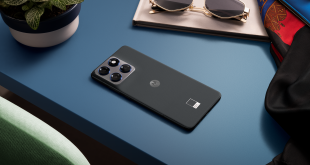 Moto Edge 70 lying face down on a blue desk beside sunglasses, a notebook, and a plant, showing its slim profile and triple‑camera module.