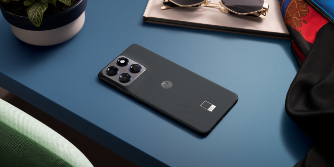 Moto Edge 70 lying face down on a blue desk beside sunglasses, a notebook, and a plant, showing its slim profile and triple‑camera module.