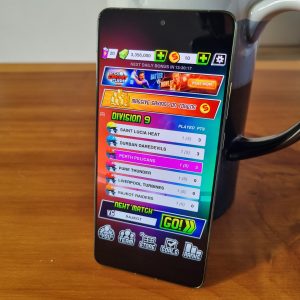 Moto Edge 70 upright on a wooden table in front of a mug, displaying the bright menu from Super League with colourful icons and text.