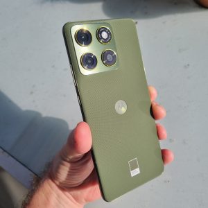 Hand holding a green Moto Edge 70 outdoors, highlighting the textured back, Motorola logo, and square camera module with three lenses and gold accents.